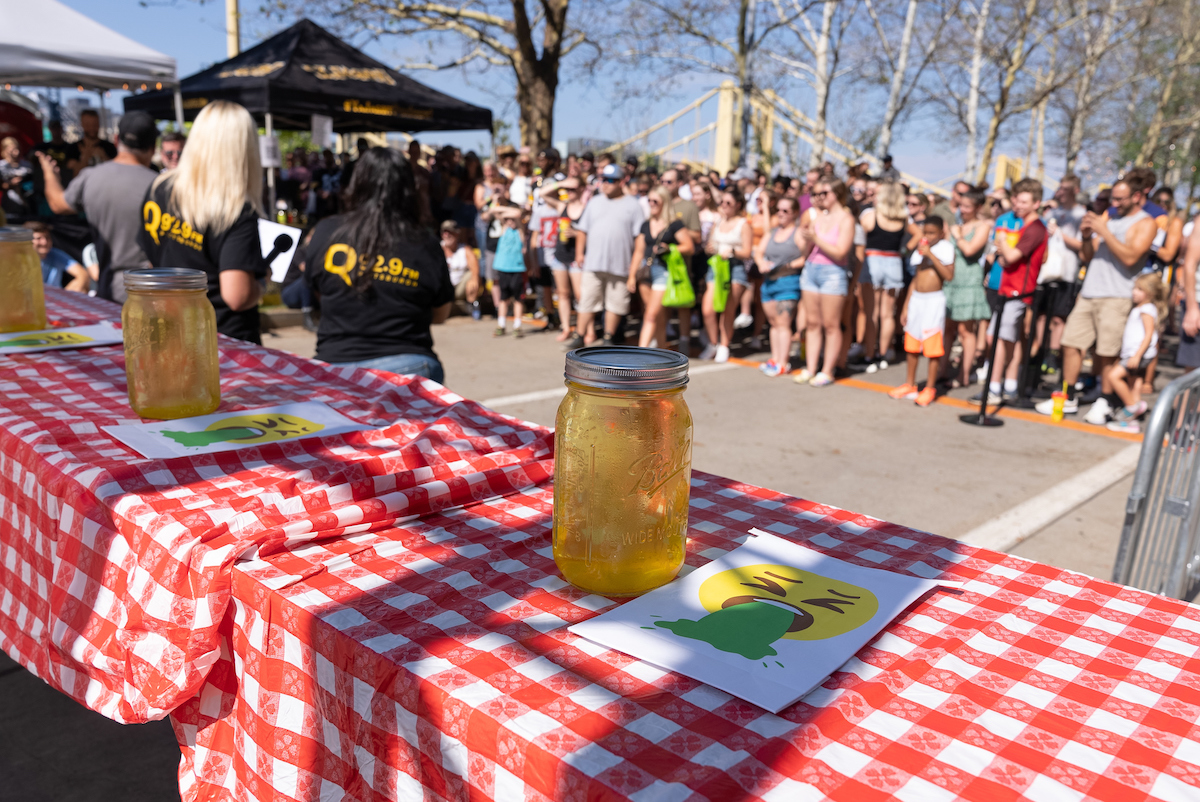 A crowd stands facing a table with jars of pickle juice on it.