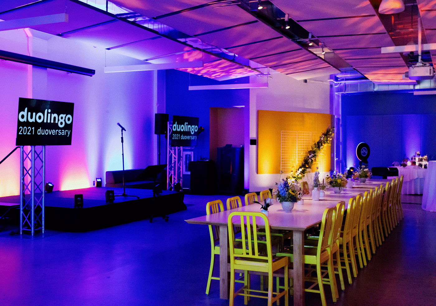 A stage with two screens sits in a room with tables and yellow chairs