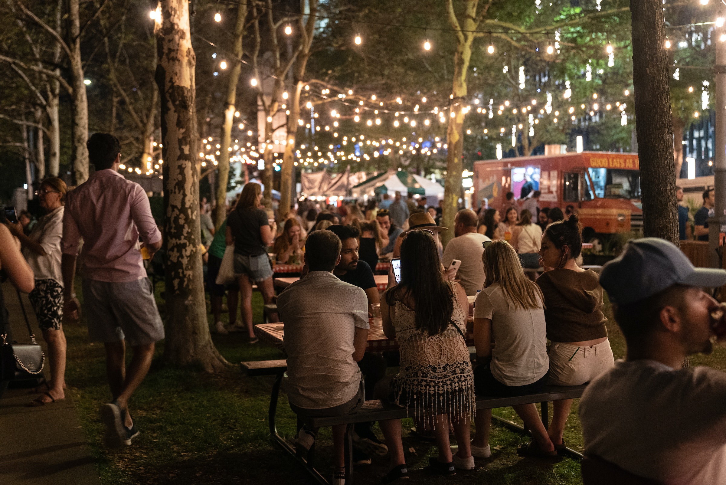 People Sit At Picnic Tables Below String Lights