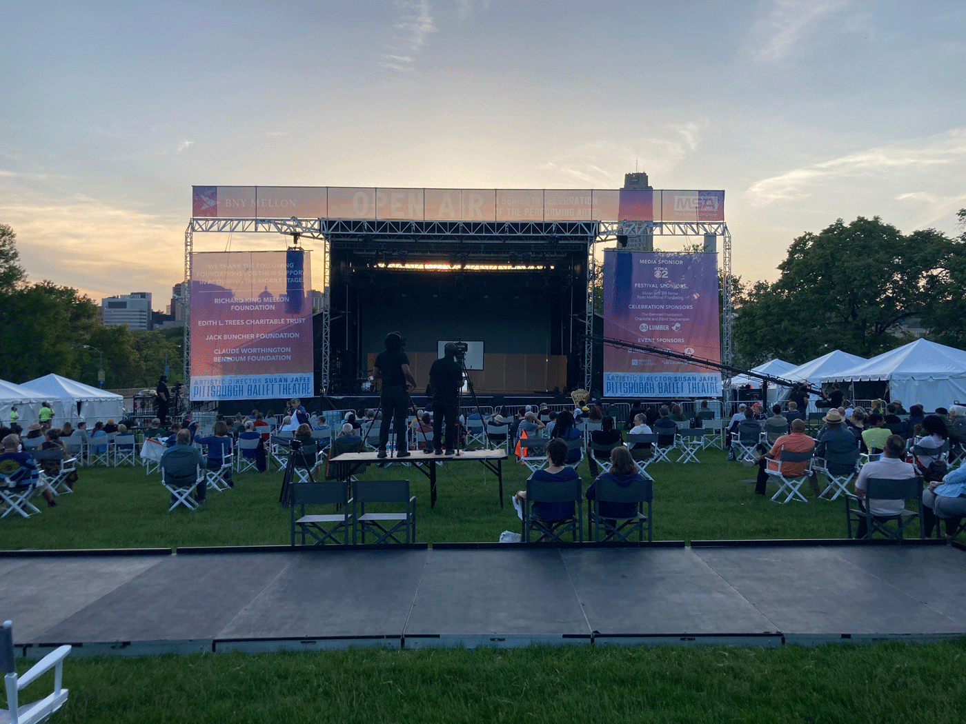 A Crowd Sits On A Lawn Looking At An Outdoor Stage