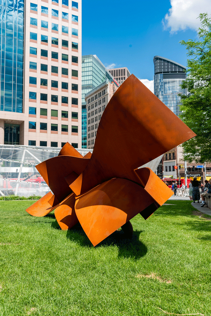 A geometric, orange sculpture sits on a lawn in the city
