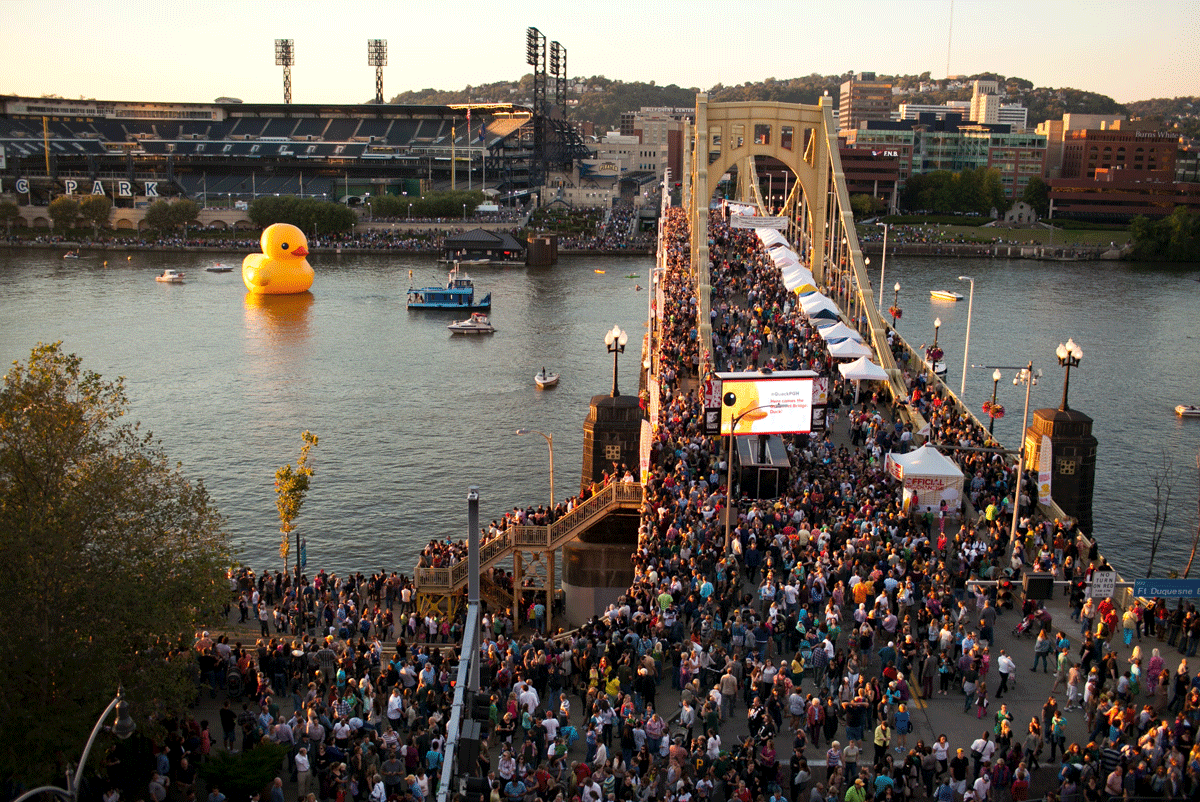 A crowd is gathered on a bridge looking at a giant duck in a river