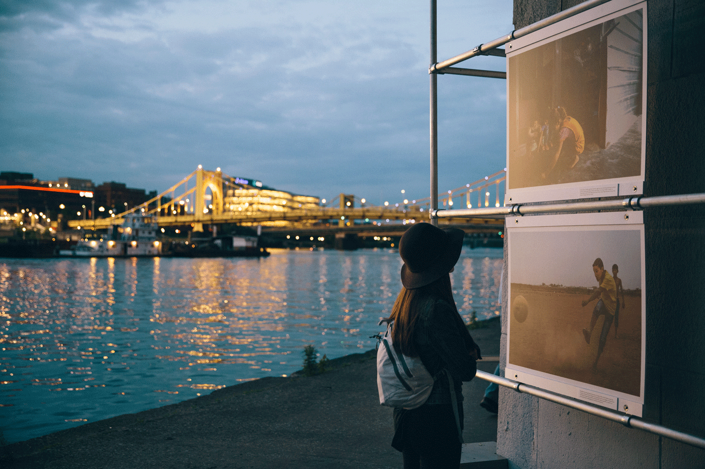 A person stands with a bridge and river behind them, looking at hanging photos